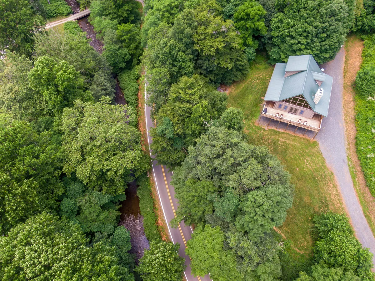 A wide view of the house set amid trees, showcasing the winding road, the flowing creek across the road, and the surrounding natural landscape.