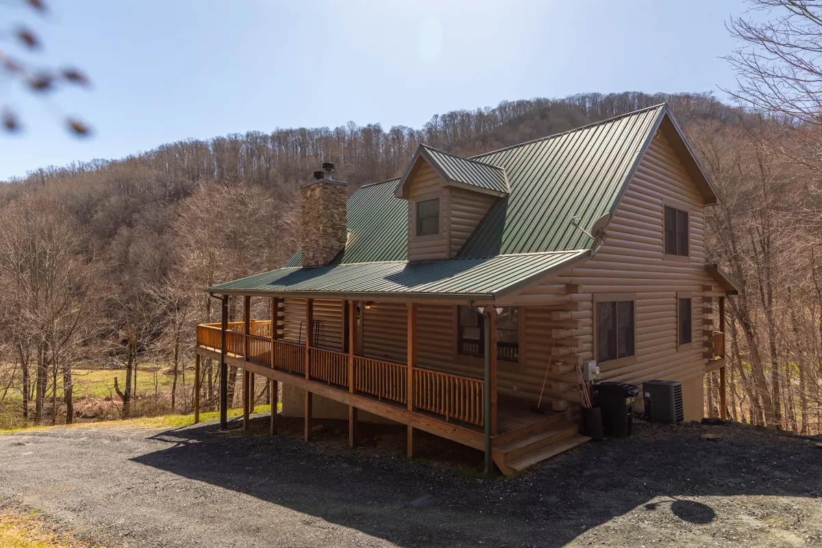 A classic log cabin exterior with a green metal roof, wraparound porch, and a stone chimney, surrounded by lush natural scenery. The porch and yard are set for outdoor lounging and family gatherings, encapsulating the spirit of mountain adventures.
