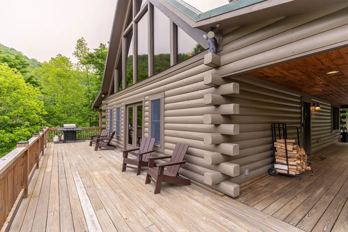 This exterior shot captures the welcoming log cabin structure with large windows, an expansive outdoor deck, wooden chairs, and lush greenery surrounding the property, inviting guests to relax, socialize, or enjoy nature walks.