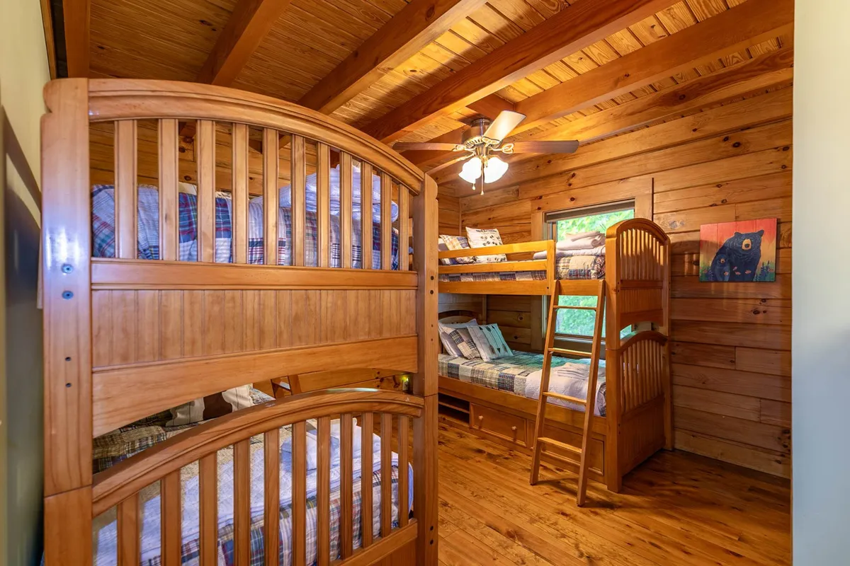 A cheerful bunk room featuring wooden bunk beds, a ceiling fan, and colorful artwork on the wall. The room exudes a playful, welcoming vibe, perfect for children and teens to bond and enjoy sleepovers.