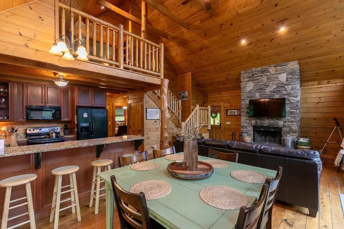 A wide-angle view of the central living space reveals the stone fireplace, a farmhouse dining table, visible kitchen counter, and rustic wooden beams. The arrangement encourages group interaction—ideal for board games, shared meals, and cozy relaxation.
