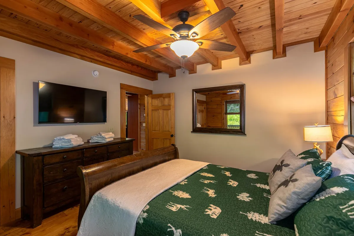 A spacious, inviting bedroom with a king-sized bed, wooden ceiling beams, a television, wall mirror, and a dresser. Natural light fills the room, creating a serene and restful atmosphere ideal for adults seeking privacy and comfort.