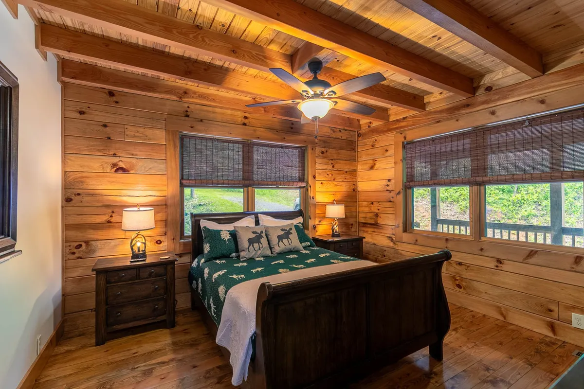 A cozy queen bedroom with a wooden bed frame, decorative pillows, table lamps, and large windows offering views of the outdoors. The room is bathed in natural light, creating a calm and welcoming space for guests.