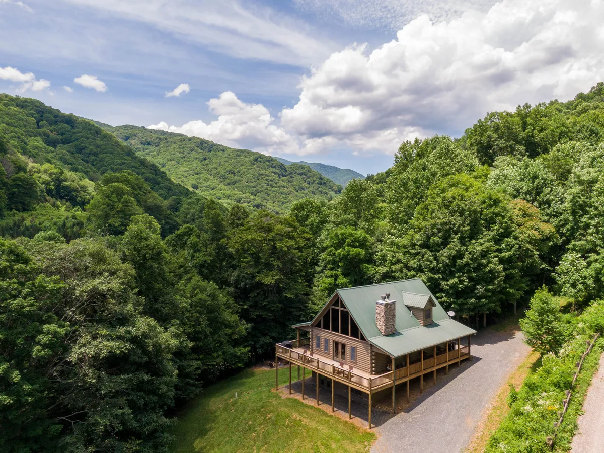 The image showcases a classic log cabin nestled among lush trees with a stunning mountain backdrop. The cabin sits at the end of a gravel driveway, inviting guests to enjoy the peaceful, natural setting. The porch hints at relaxing mornings, and the open yard is perfect for gathering or playing. The entire scene radiates tranquility and a sense of escape into the mountains.