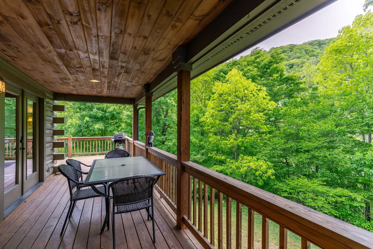 A close-up of the wooden deck surrounded by lush greenery, featuring a glass table and black chairs. The tranquil setting highlights the opportunity for outdoor dining, lounging, or simply enjoying nature with pets nearby.