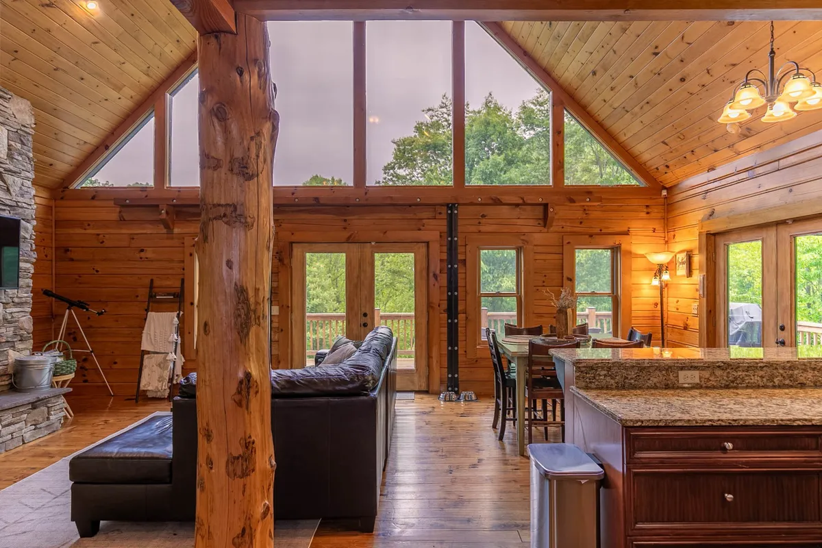 This image showcases the heart of the lodge: a spacious living area with large windows framing lush greenery, a dramatic floor-to-ceiling stone fireplace, rustic wooden beams, a granite kitchen countertop, and a cozy sofa. The open-concept layout visually connects the living room, kitchen, and dining spaces, highlighting natural light and inviting communal gatherings.