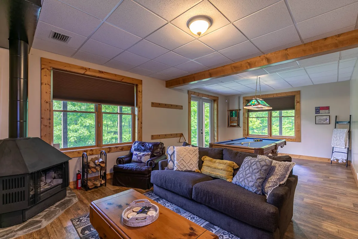 A spacious entertainment area is shown with a pool table in the foreground, a cozy sofa, a warm fireplace, and large windows that let in plenty of natural light. The space feels both lively and relaxing, perfect for gathering with friends or family.
