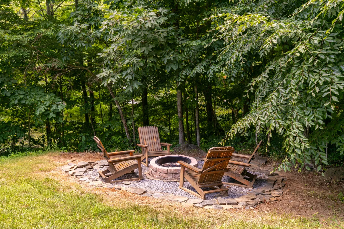 This image features a cozy outdoor fire pit surrounded by wooden chairs nestled among trees, creating an inviting communal space ideal for gathering, roasting marshmallows, and enjoying nature.