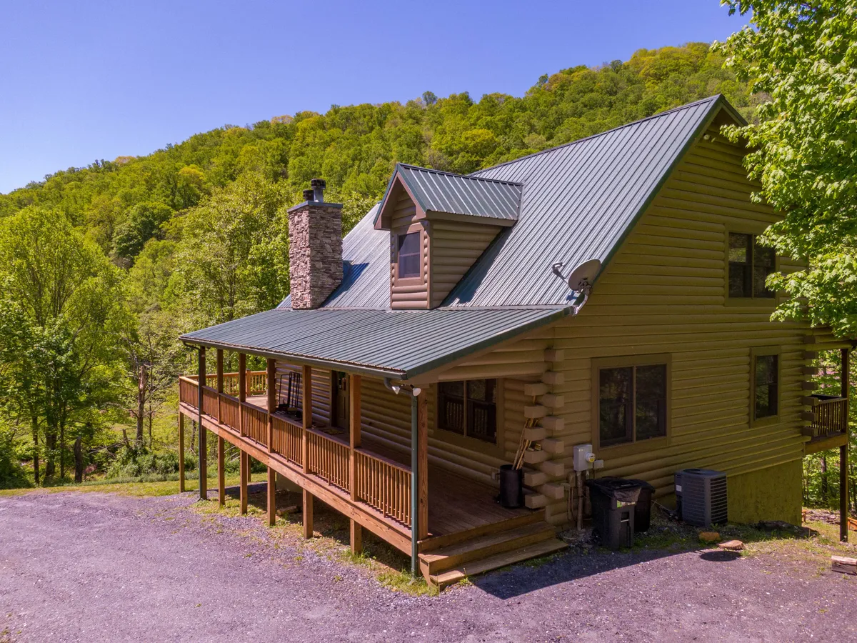 This image features the log cabin's classic exterior, highlighted by a green metal roof, stone chimney, and a welcoming wraparound porch. The porch opens out into spacious, natural surroundings, hinting at ample space for pets to play and families to unwind together outside.